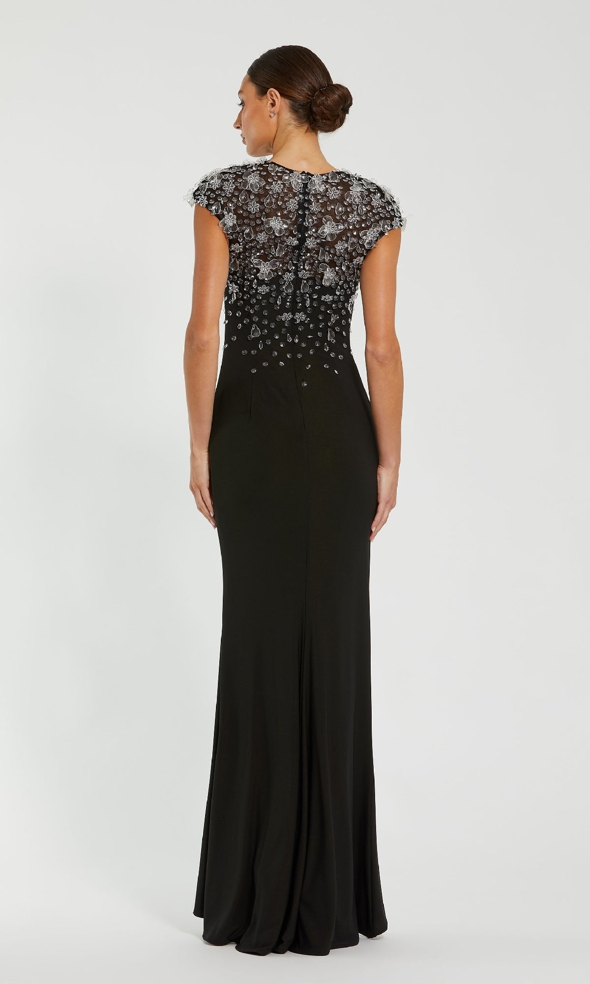 A woman, hair in a neat bun, faces away wearing the Mac Duggal 44081 Cap Sleeve Black Formal Dress—a floor-length gown with sheer embellished lace back and cap sleeves.